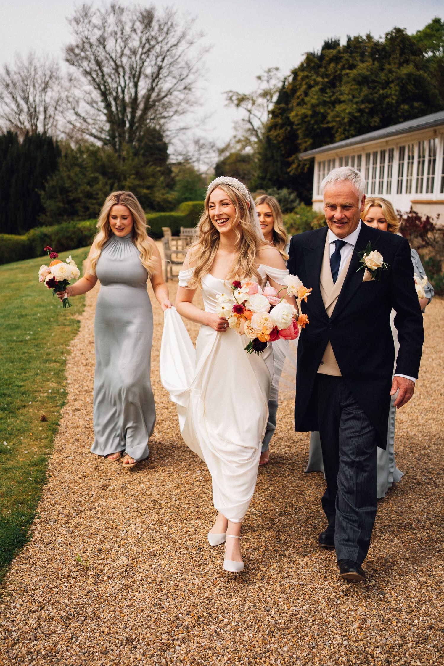 A wedding party walks and laughs together along a gravel path outside on a sunny day.