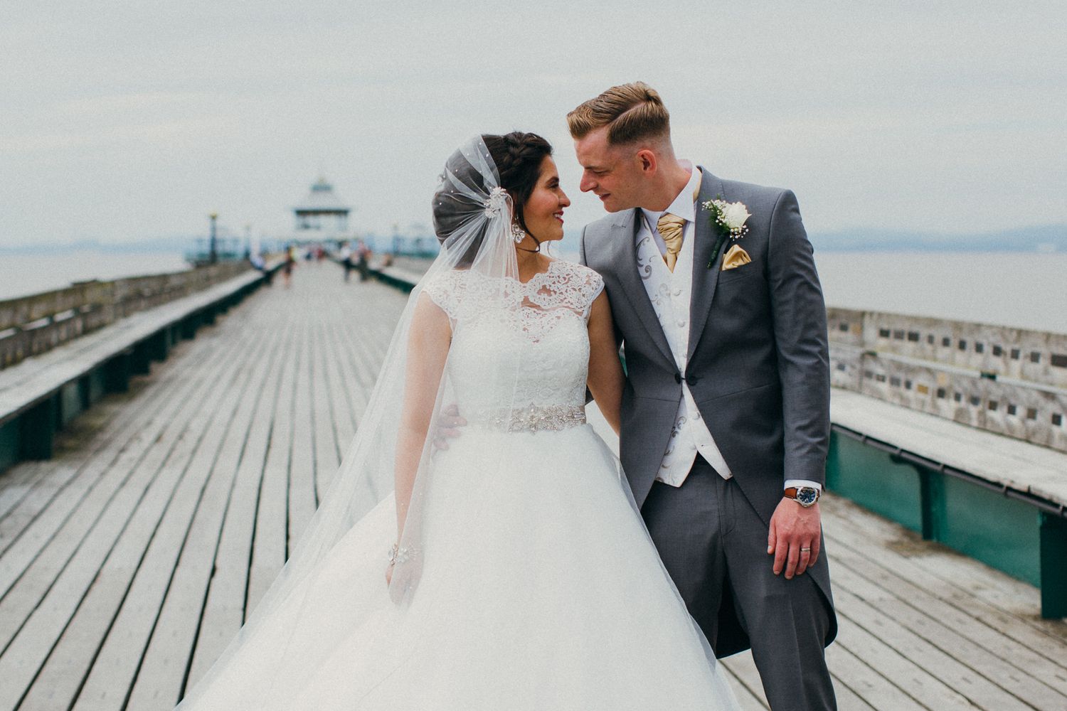 A couple in wedding attire walks along a wooden boardwalk near the ocean on their special day.