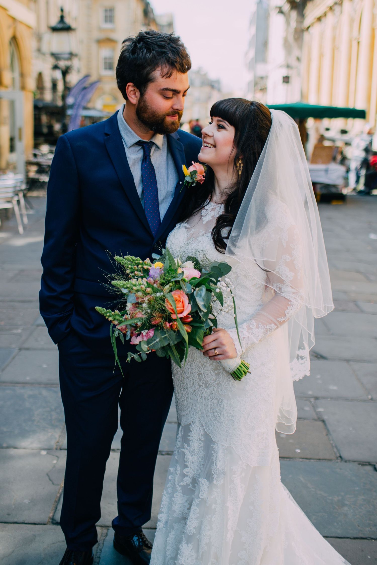 Newlywed couple sharing intimate moments outdoors with bride holding floral bouquet against urban backdrop.