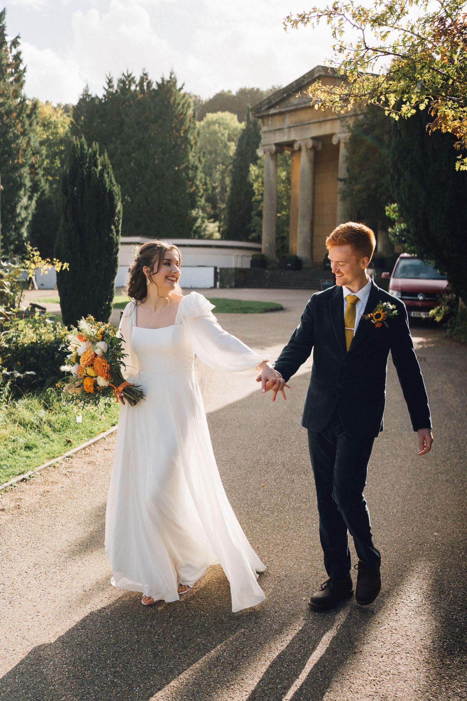 A couple in wedding attire dance and twirl together on a sunlit path outside a stone building.