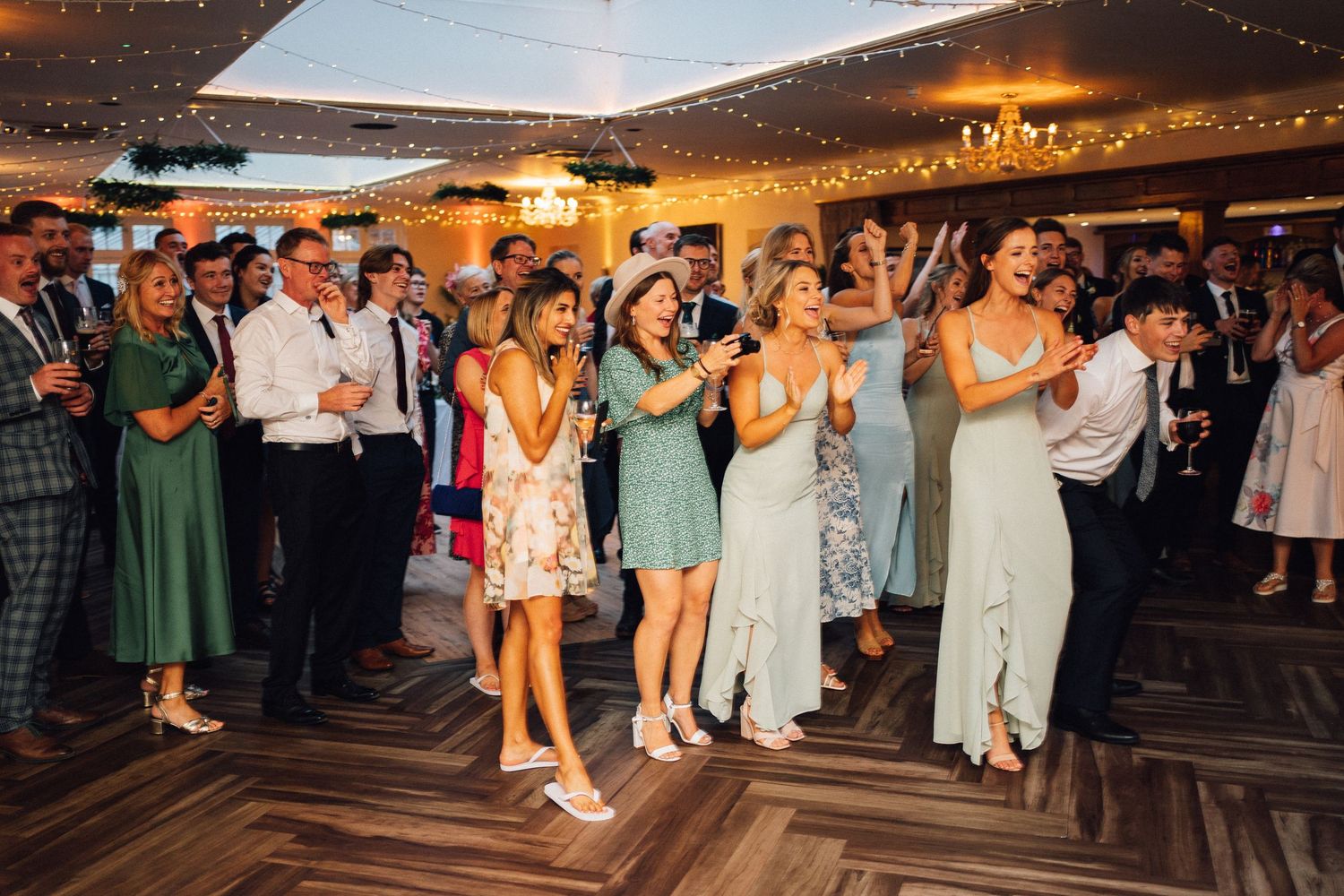 Wedding guests dance and celebrate on wooden floor under string lights at evening reception.