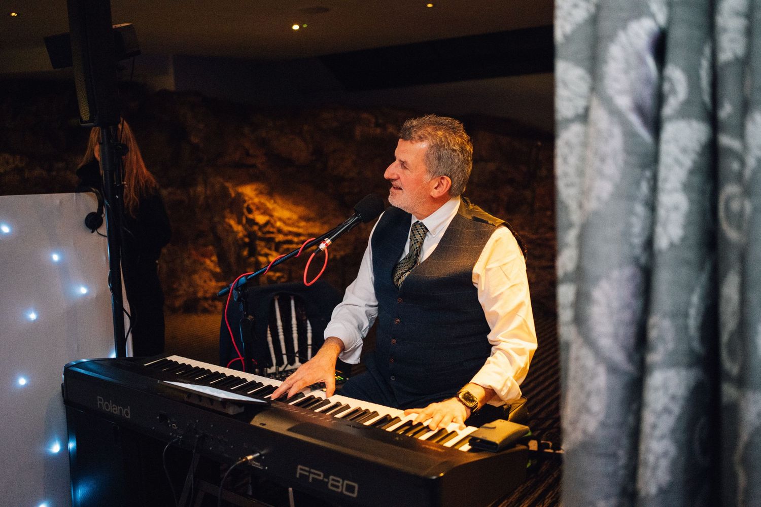 Wedding band performer playing keyboard at an evening reception with ambient lighting.
