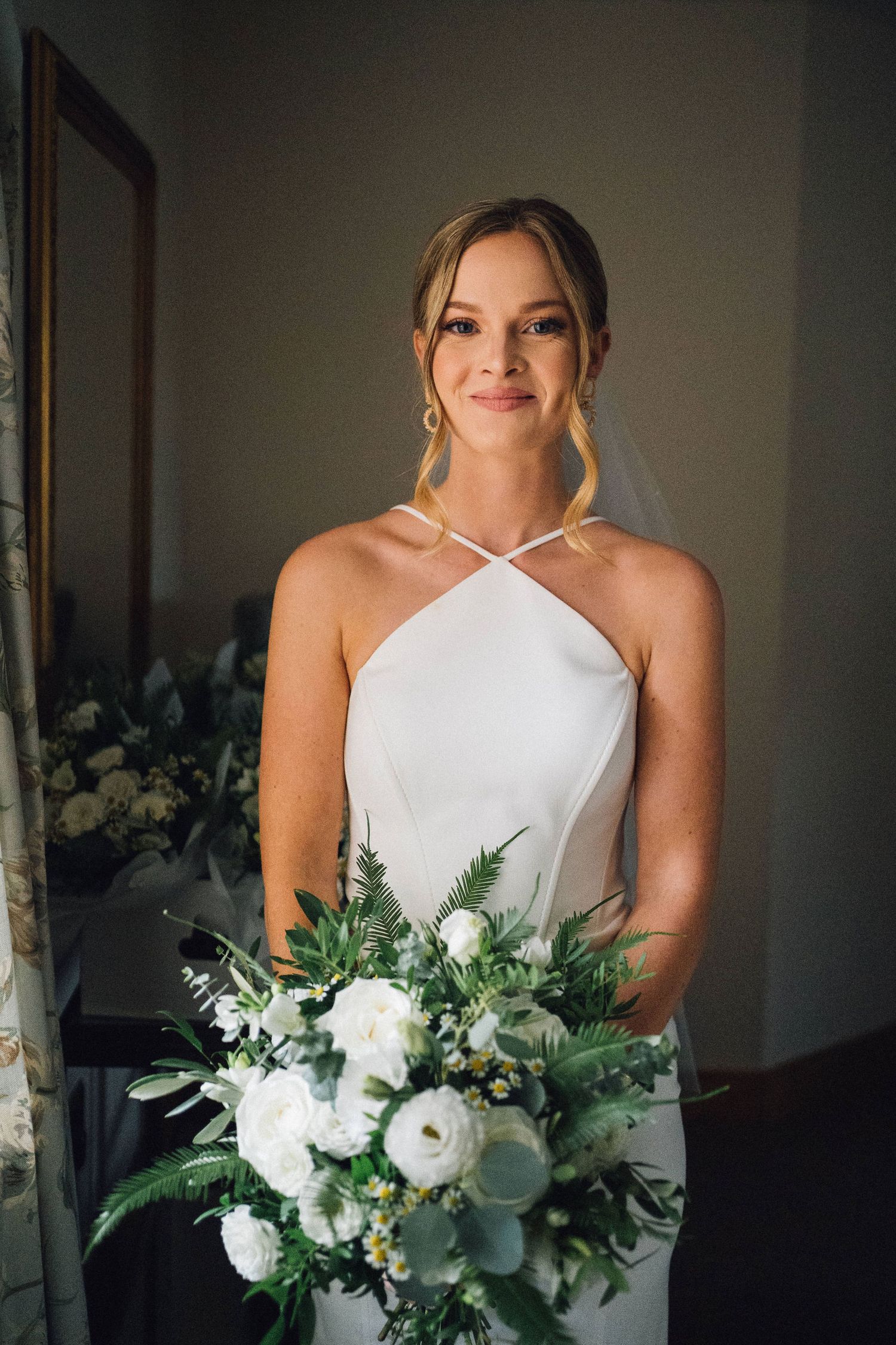 A bride in a white halter wedding dress holds a lush bouquet of white flowers and greenery in a softly lit room.