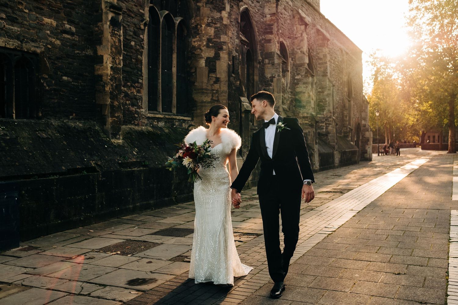 Bride and groom walking hand in hand at golden hour near a historic stone building in wedding attire.