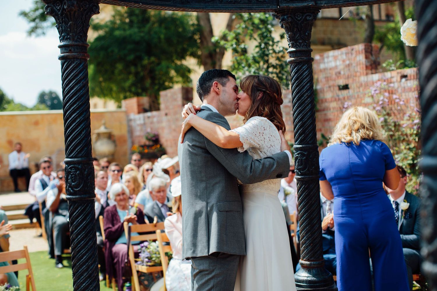 Bride and groom share their first kiss under a black iron gazebo at an outdoor wedding ceremony with seated guests.