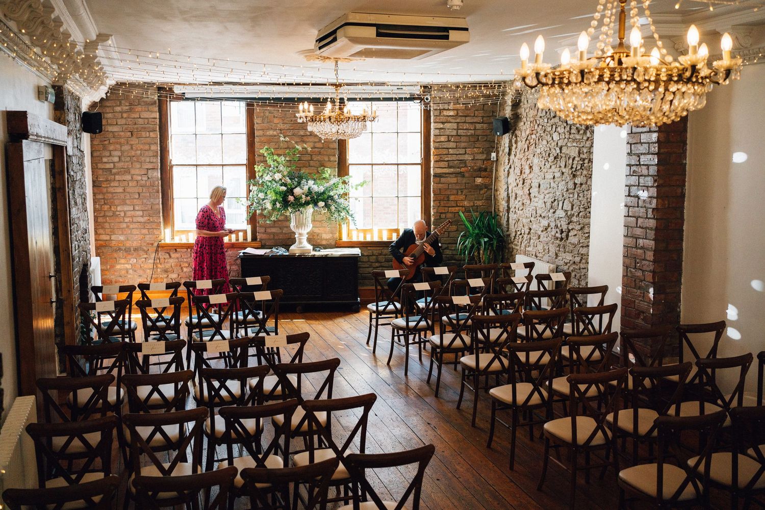 Elegant rustic wedding venue with exposed brick walls, chandeliers, and dark wooden cross-back chairs arranged in rows.