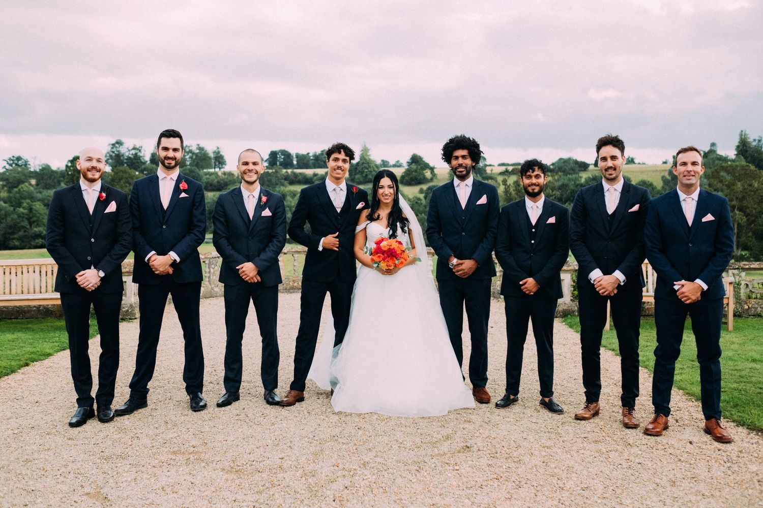 Wedding party members in black suits and pink dresses posed in a row outdoors.