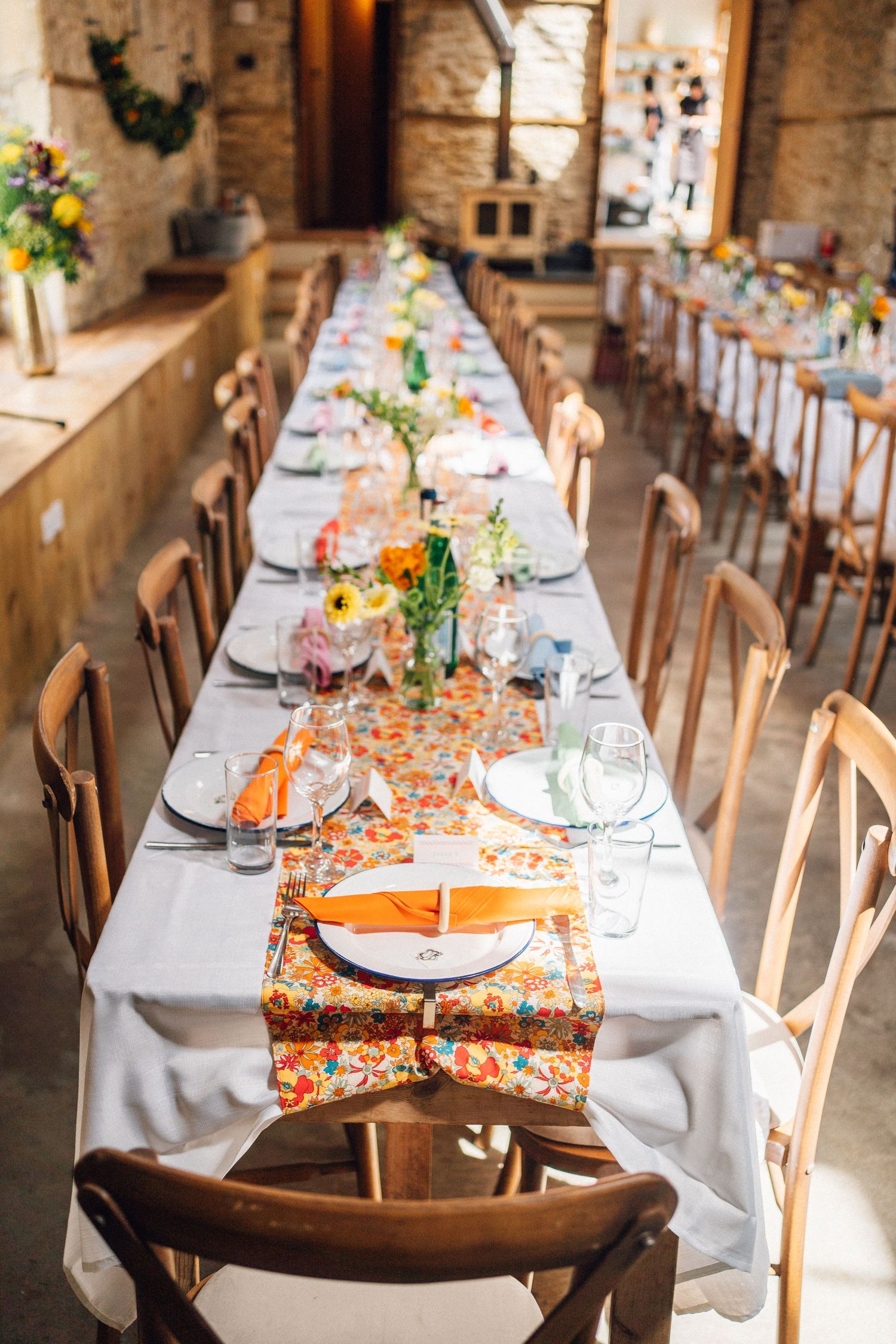 Long rustic dining tables set with white linens and colorful floral runners in a barn venue.