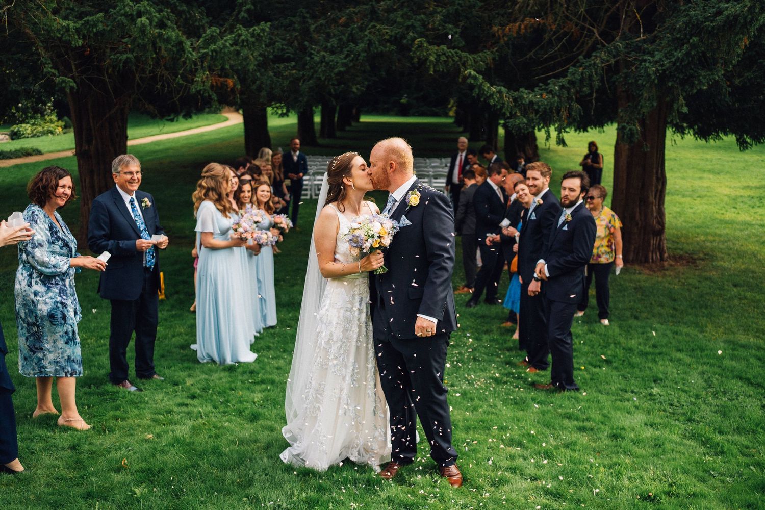 Outdoor wedding celebration with guests gathered on a sunny lawn beneath large trees.