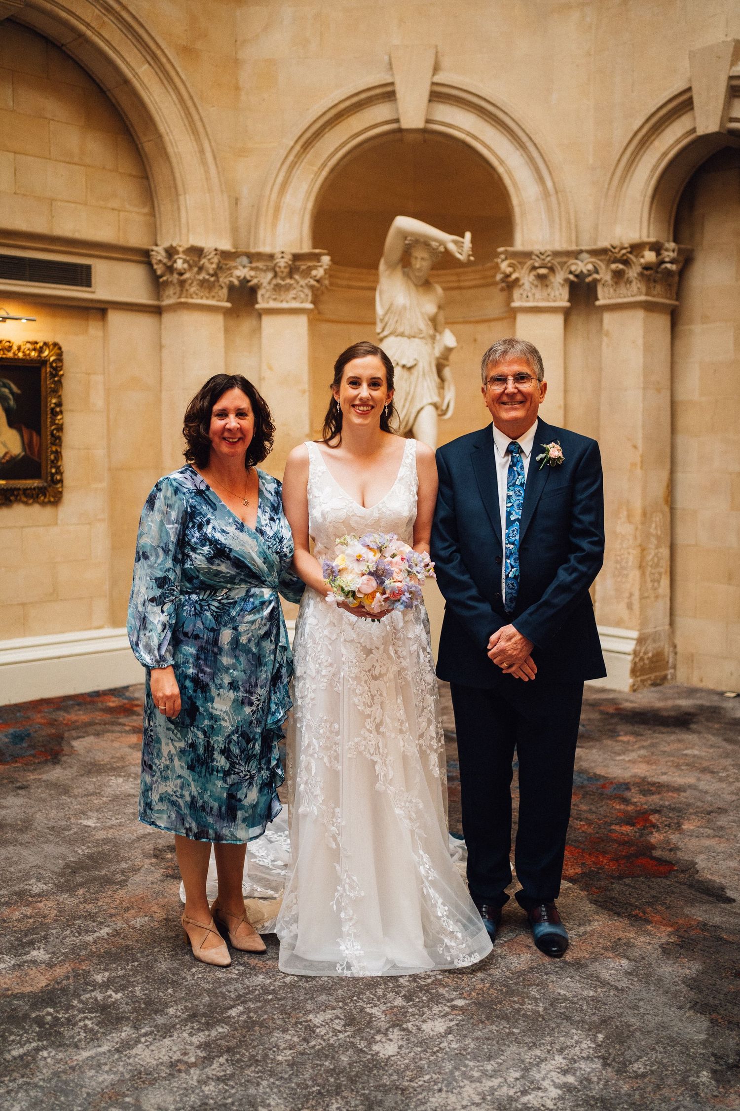 Wedding party posing in formal attire against ornate stone archways and columns.