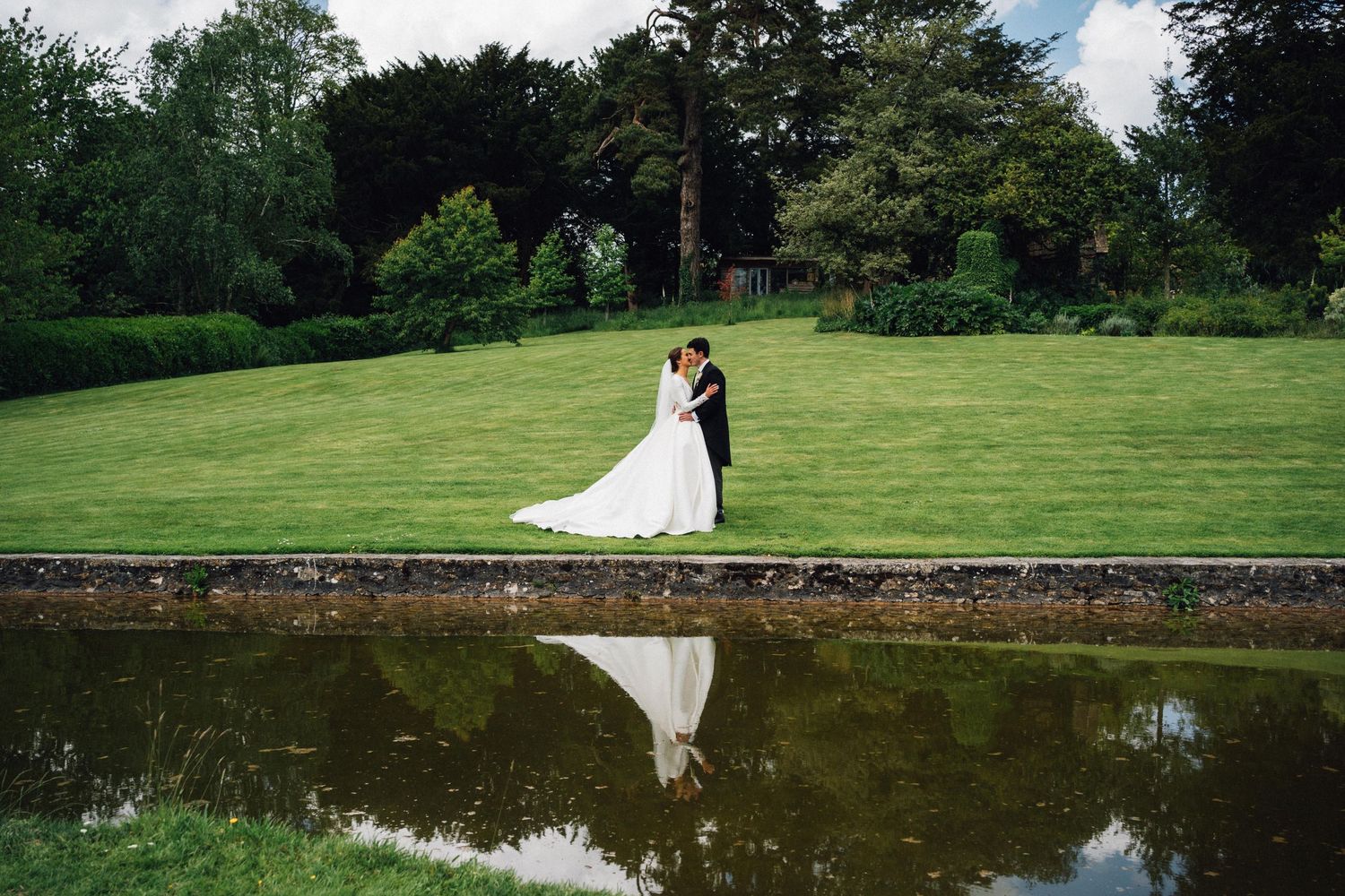 Scenic wedding venue with stone wall and reflective pond surrounded by green lawns.