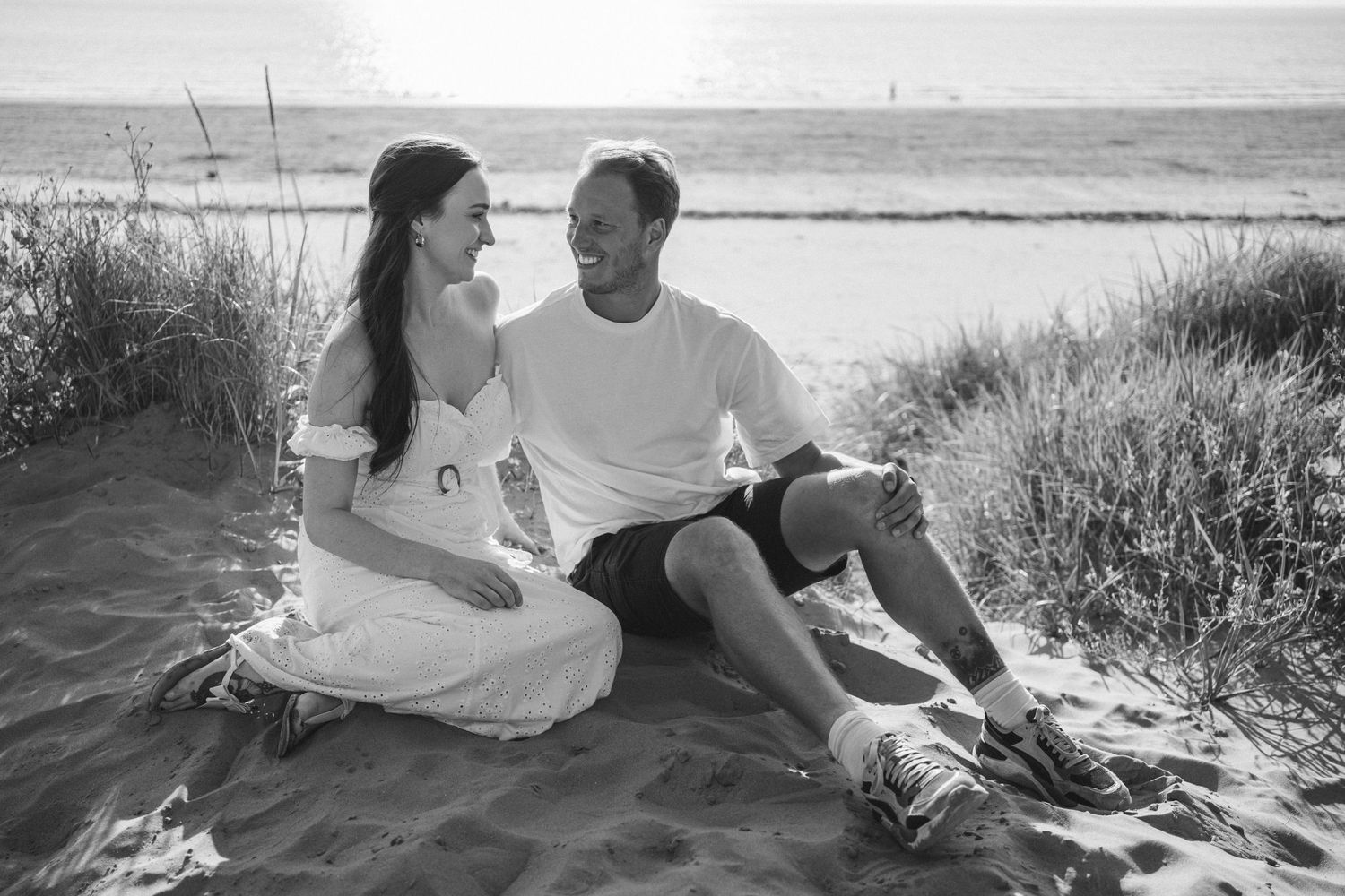 Romantic beach photoshoot with couple sitting on sandy dunes at sunset.