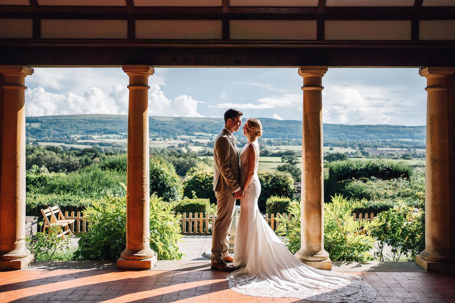 Bride and groom embrace on elegant columned terrace overlooking lush green countryside landscape on their wedding day.