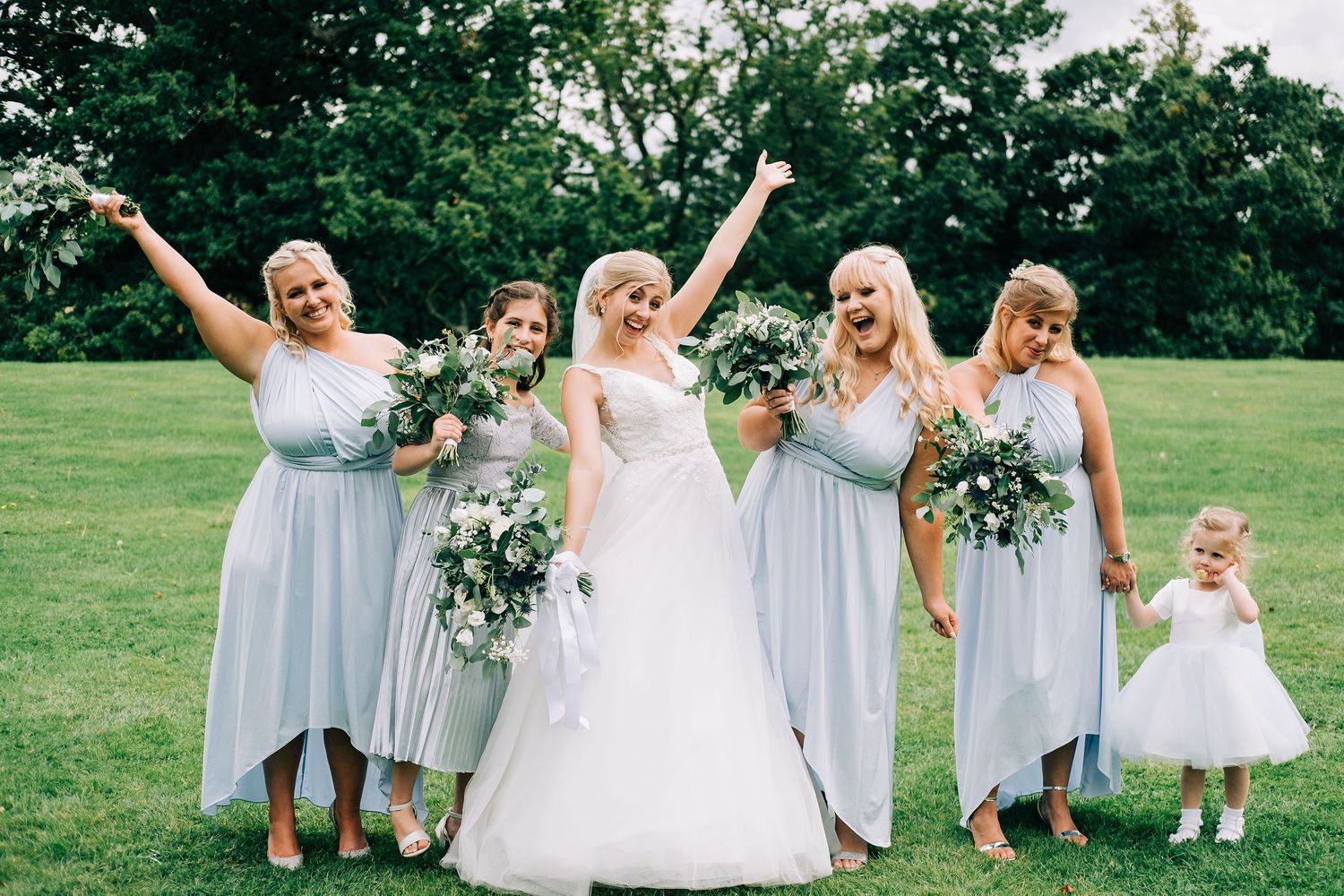 Bridal party in pale blue dresses poses joyfully with bouquets on a green lawn.
