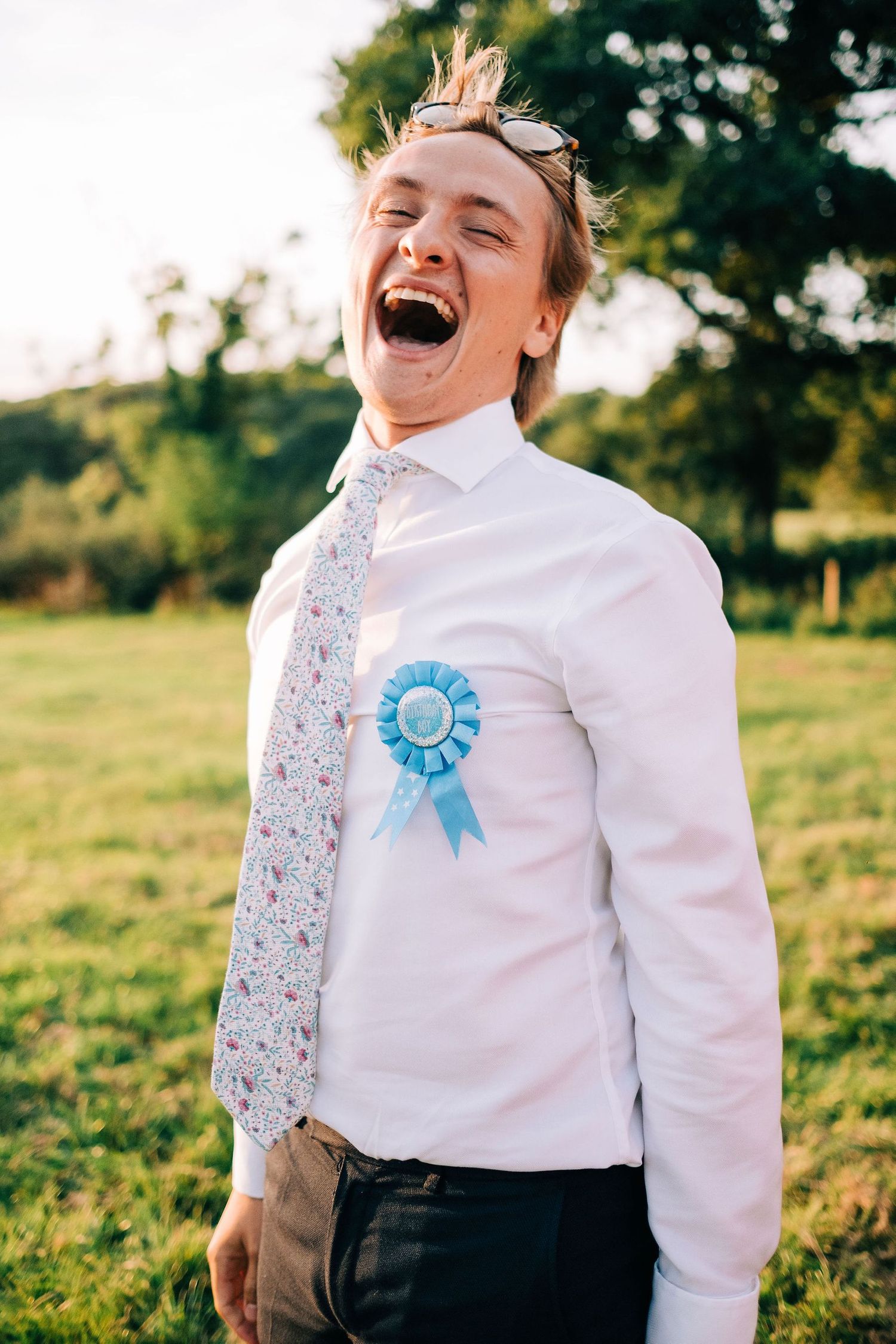 A joyful person wearing a white shirt with a blue ribbon pin laughs heartily in a sunny outdoor field.
