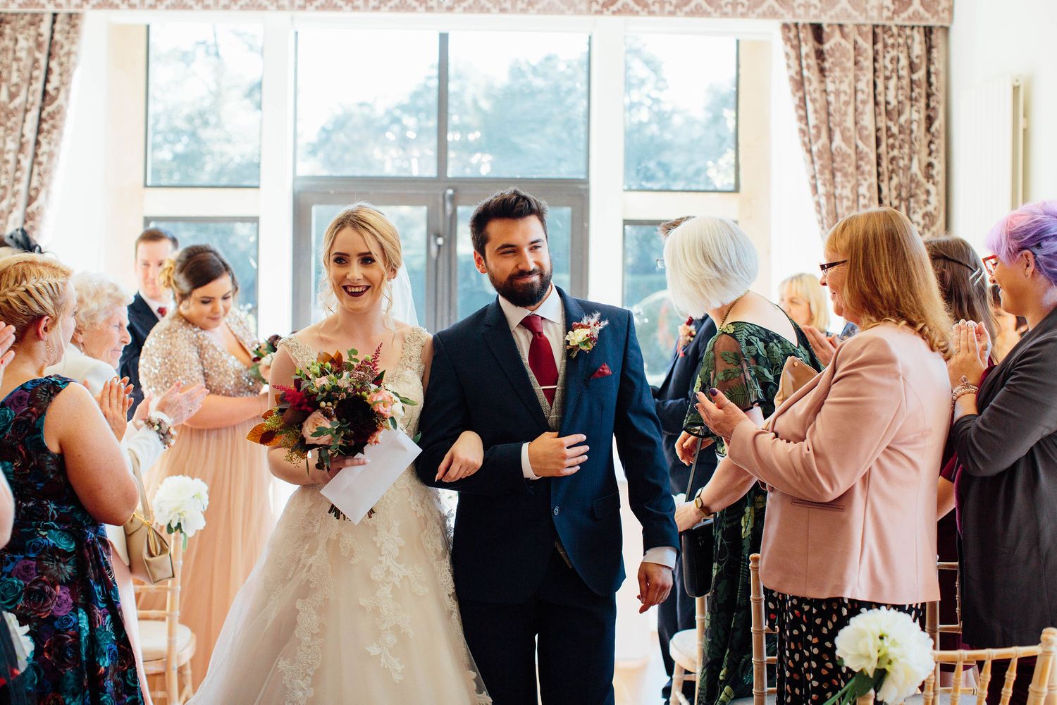 Wedding guests observe the ceremony in an elegant room with floral curtains and natural lighting.