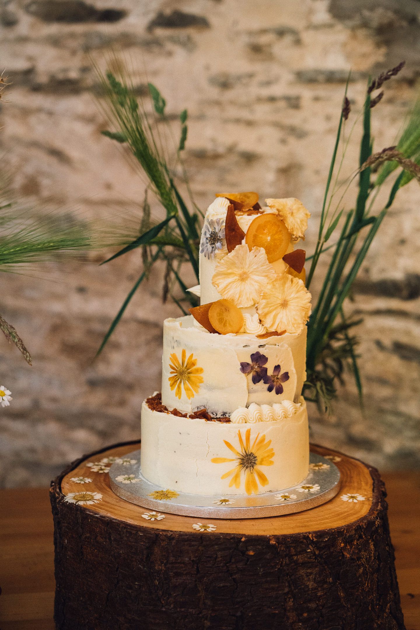 Elegant white wedding cake decorated with yellow flowers and orange accents sits atop a rustic wood slice.