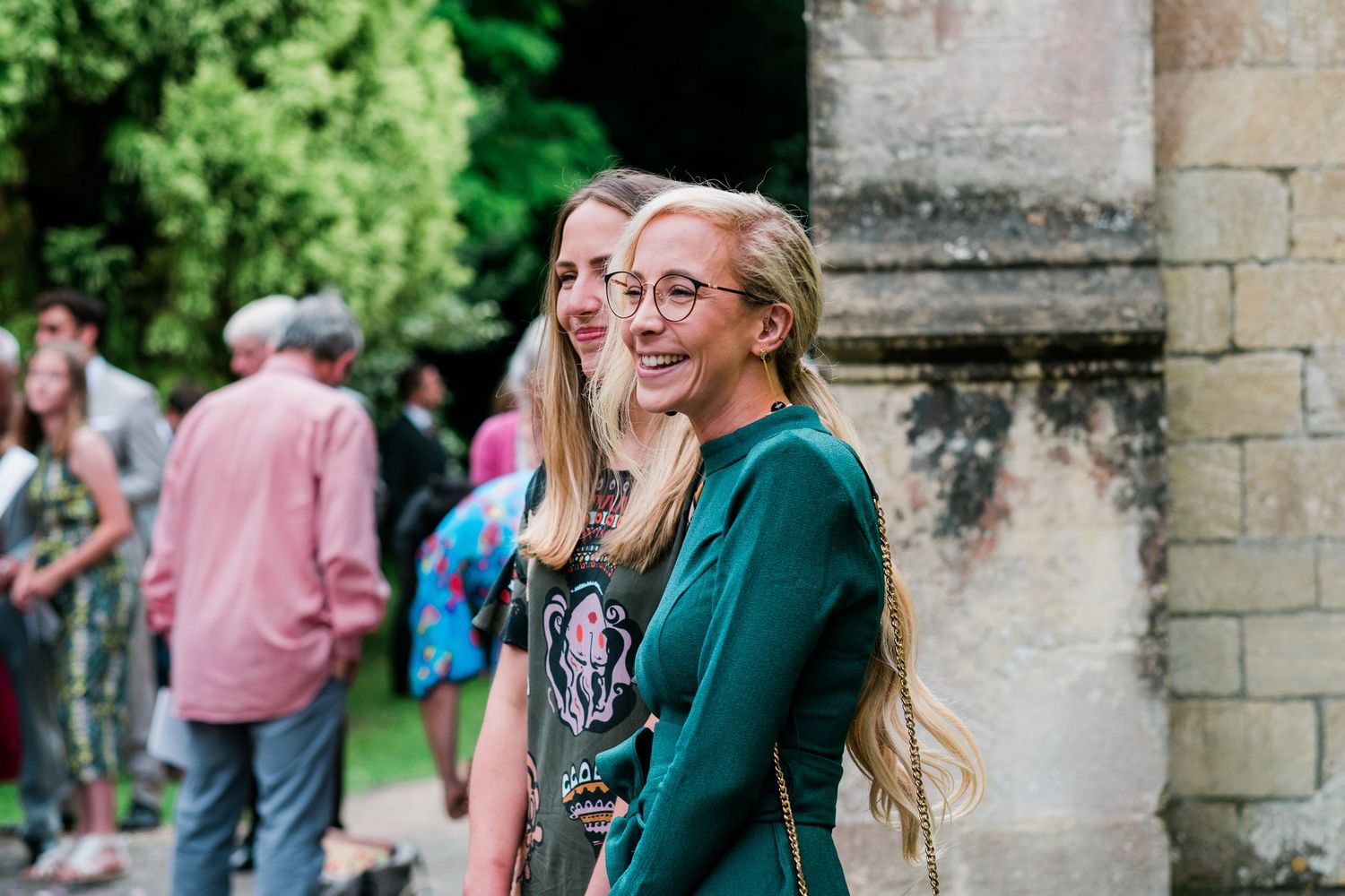 Wedding party mingles outside stone church surrounded by lush greenery.