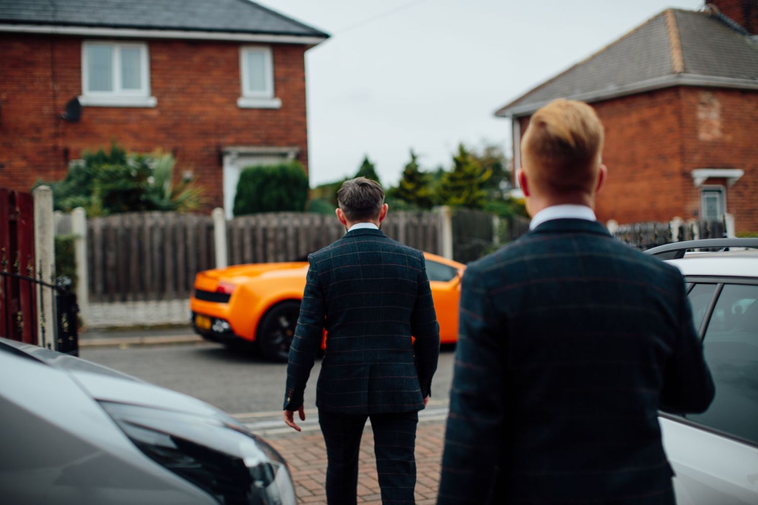 Two people in dark suits walking towards an orange sports car parked outside brick houses.