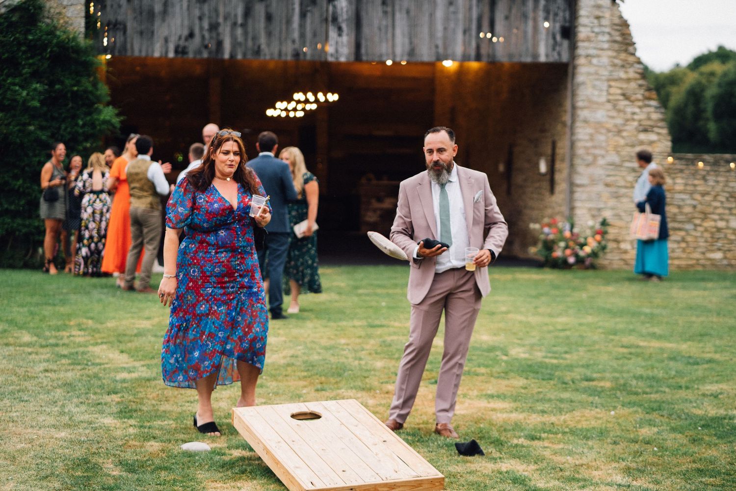 Guests playing cornhole lawn game at outdoor wedding reception near rustic barn venue.