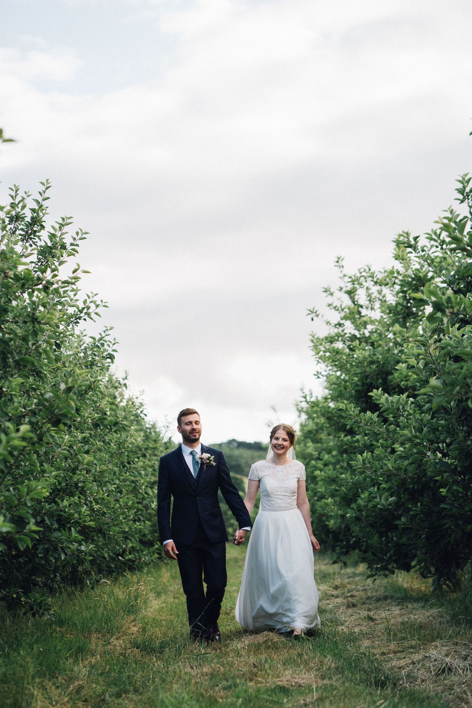 A couple walks hand in hand through an orchard path on their wedding day.