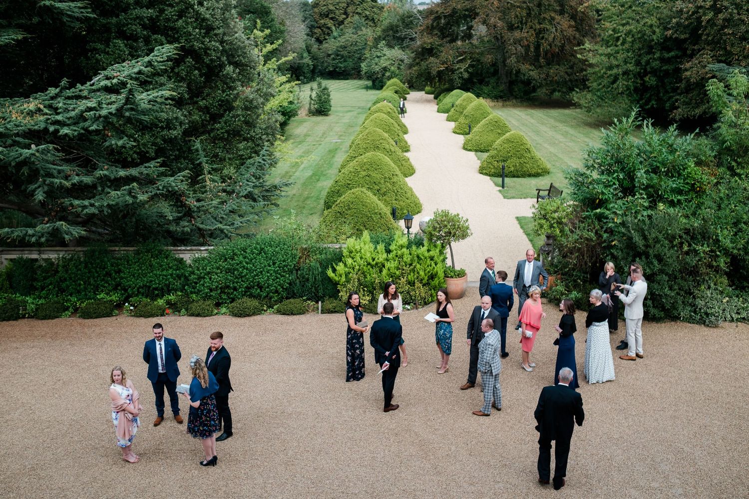Wedding guests walk along a scenic garden path surrounded by lush greenery and manicured landscaping.