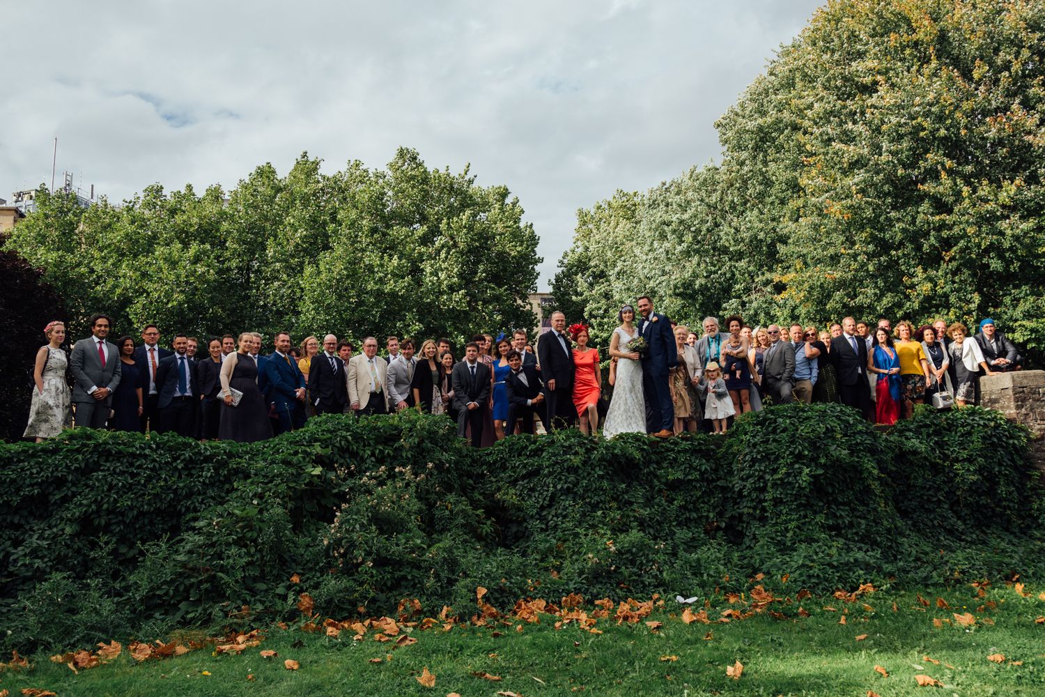 Large group of wedding guests gather for outdoor photos on a green lawn with trees in the background.