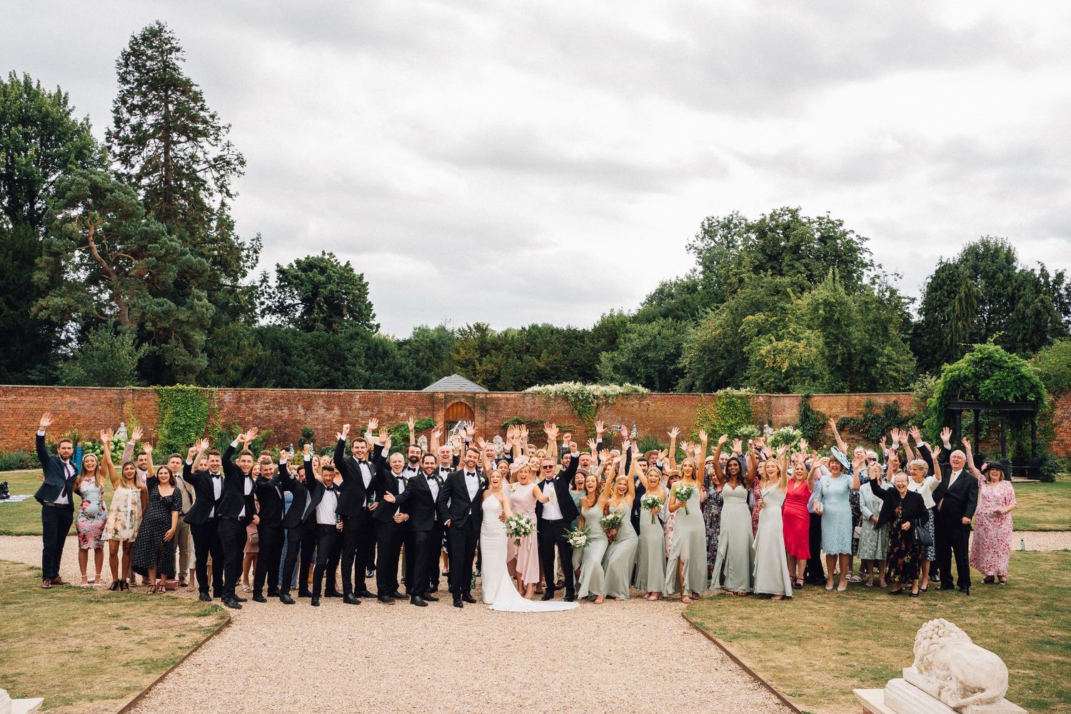 Large wedding party celebrates outdoors with raised hands against backdrop of red brick wall and green trees on cloudy day.