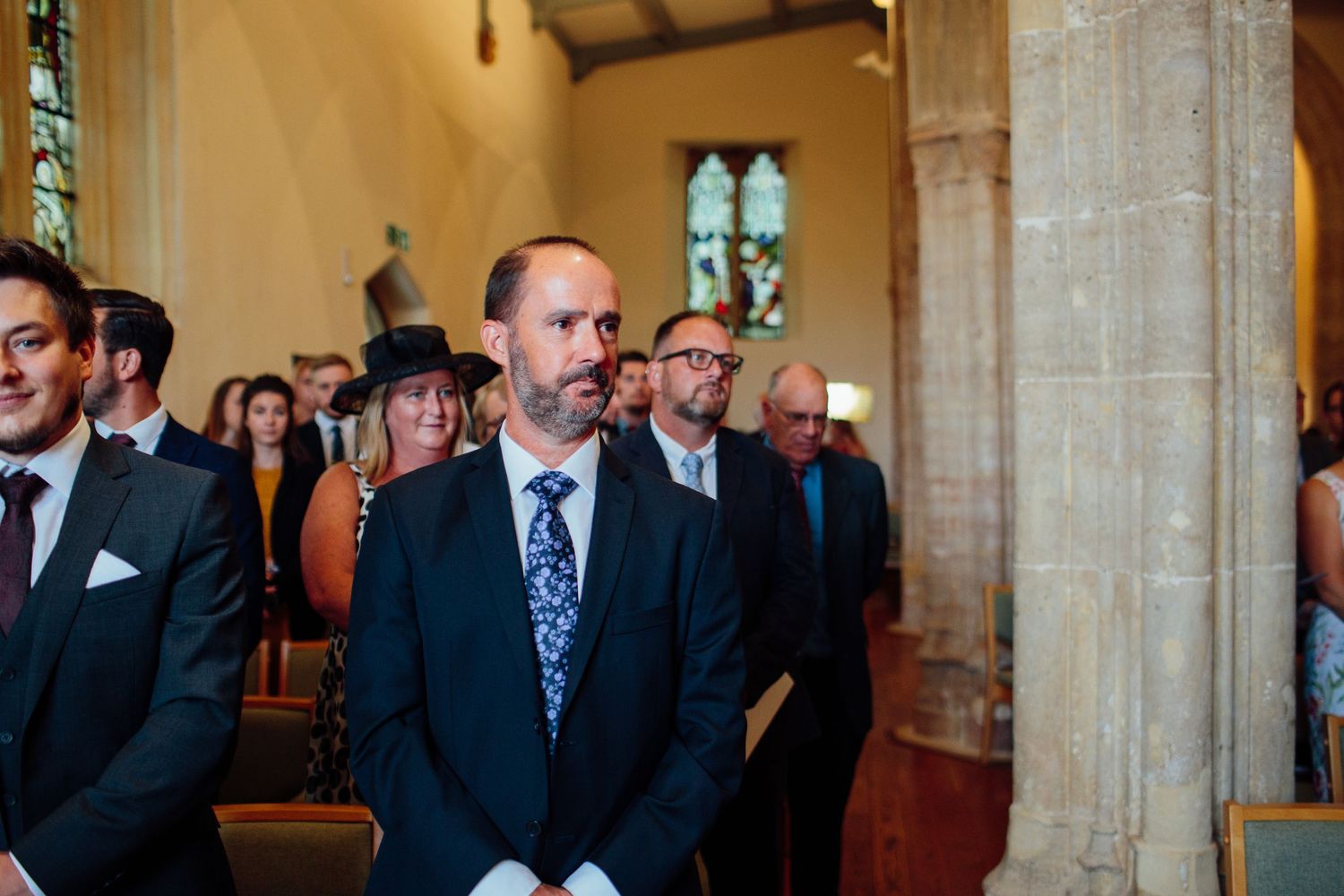 Wedding guests stand in a traditional church interior with stone columns and stained glass windows during a ceremony.