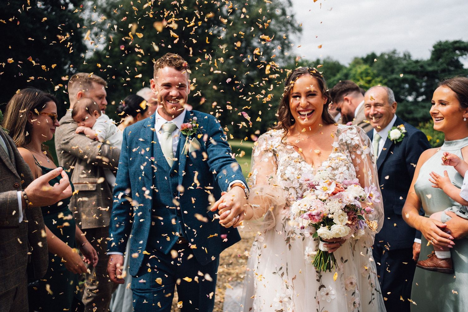 Wedding celebration with confetti being thrown over a couple in formal attire holding pink flowers outside.