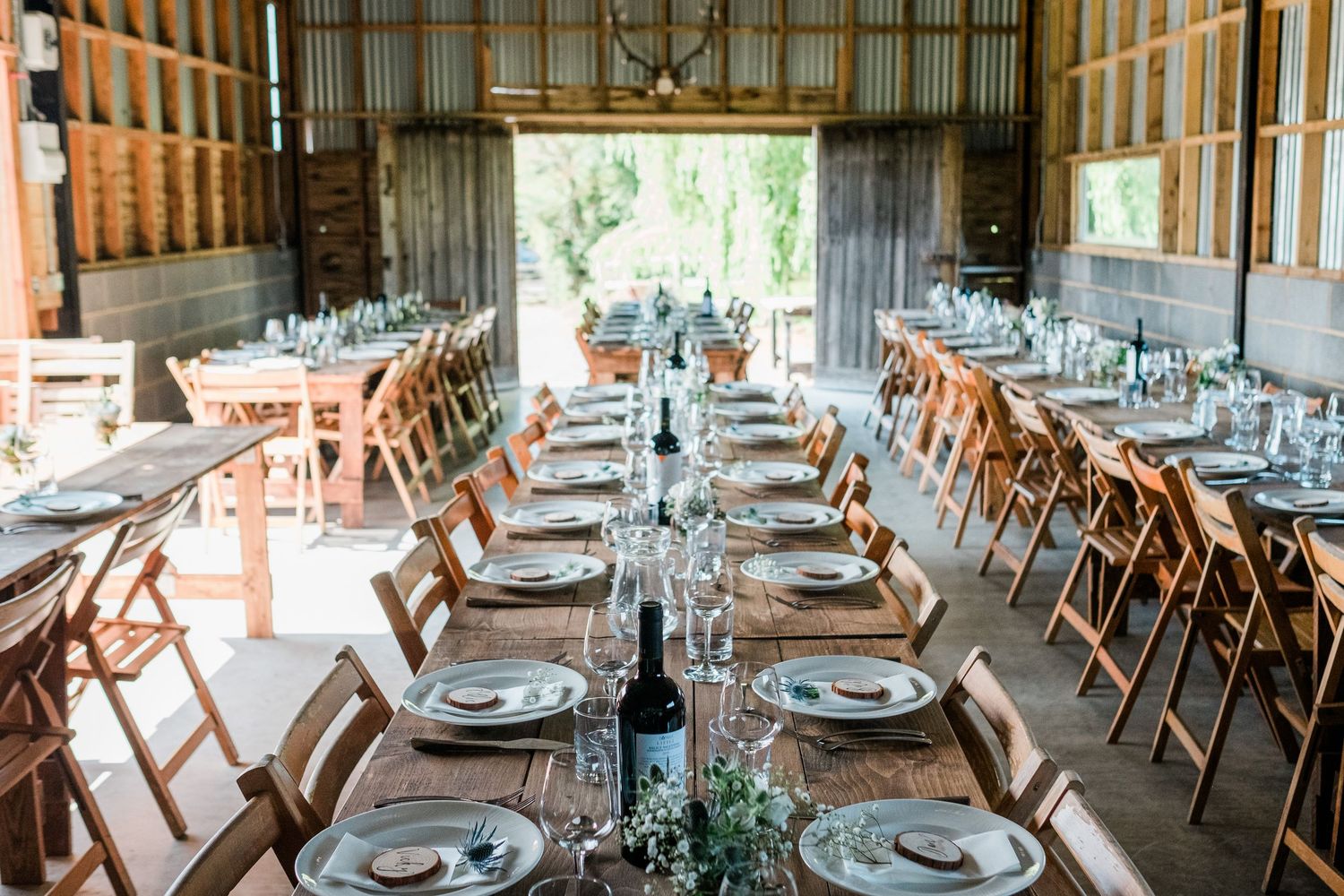 Rustic barn wedding reception venue with long wooden tables set for dinner under exposed wooden beams and natural lighting.