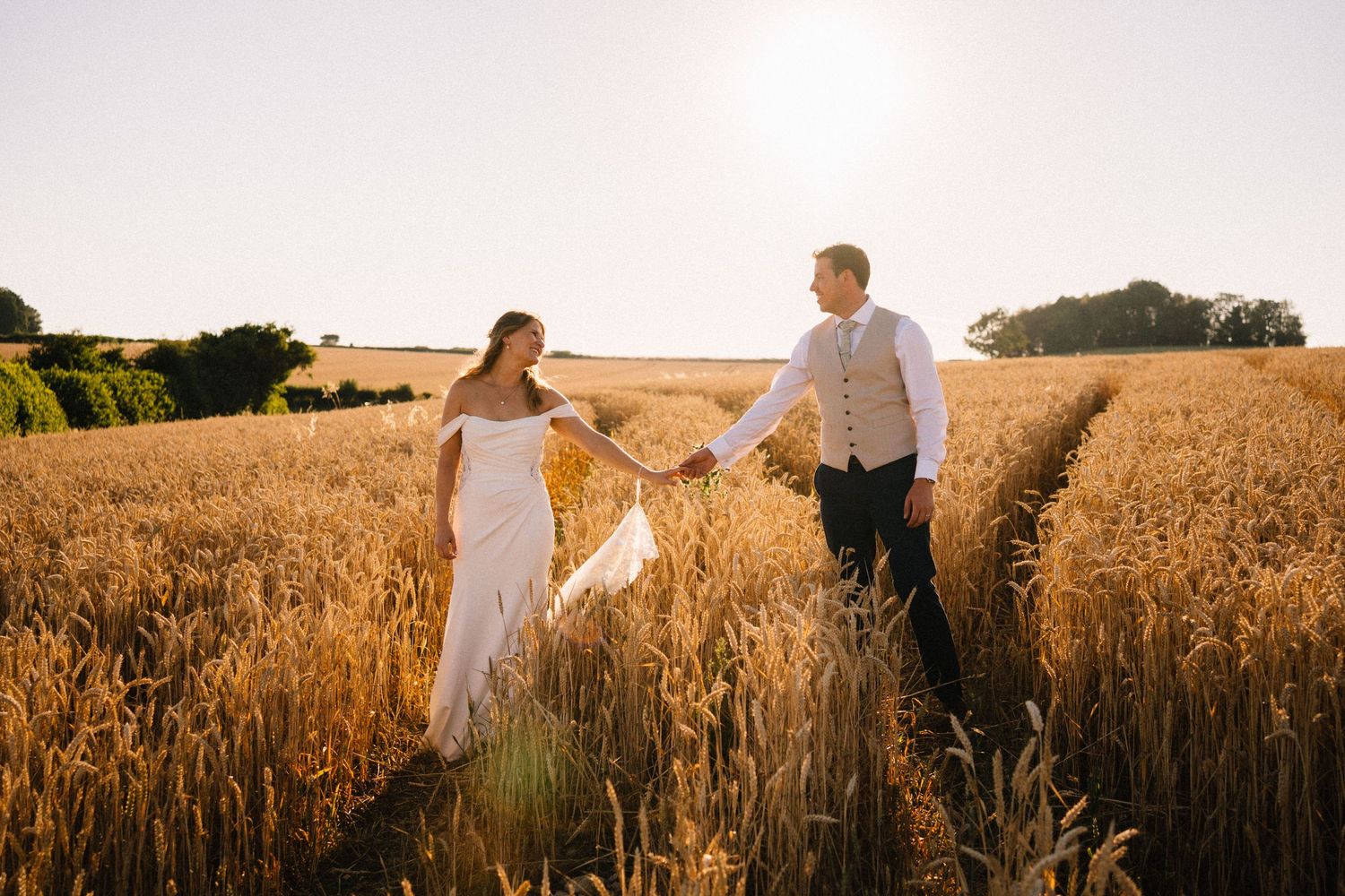 Bride and groom holding hands walking through golden wheat field at sunset in romantic outdoor wedding photo.