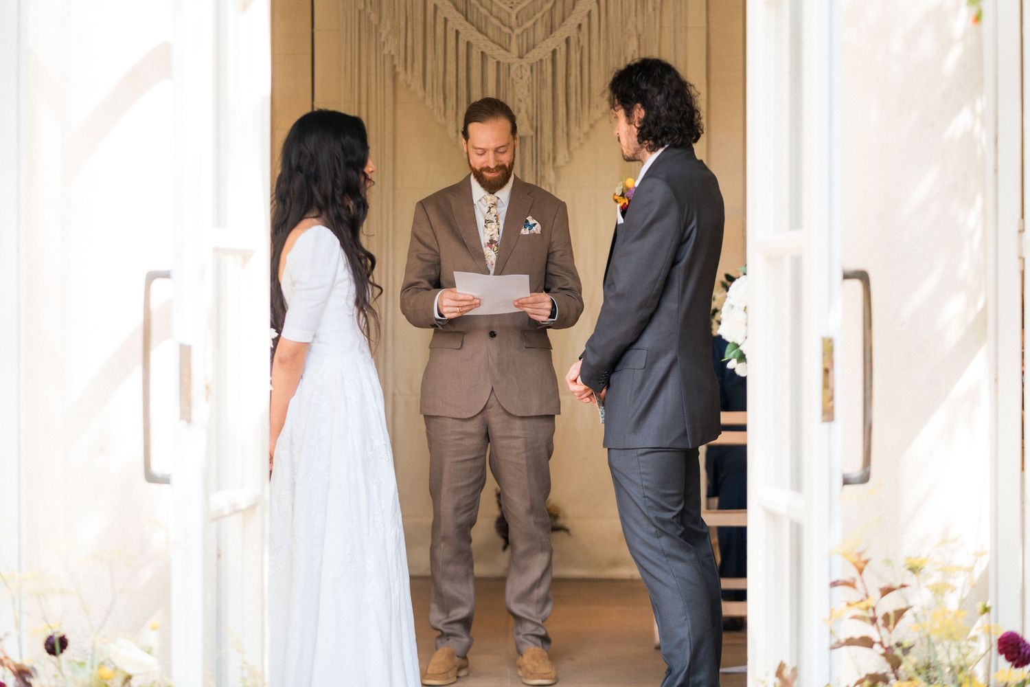 Outdoor wedding ceremony with officiant reading vows to bride in white dress and groom in gray suit.