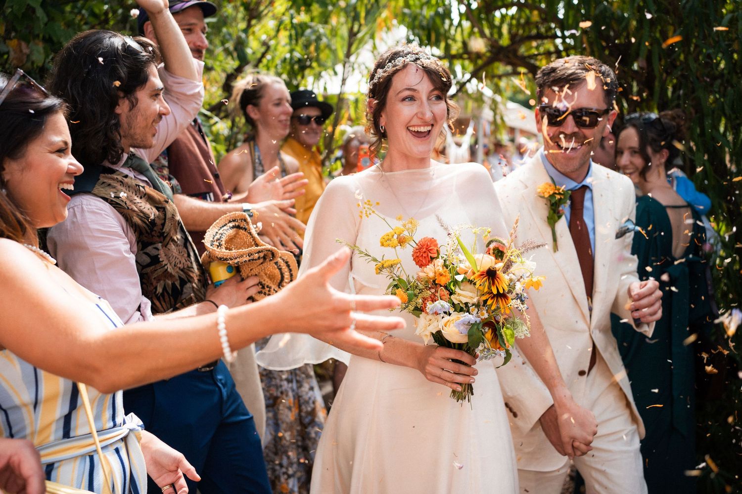 Bride and groom walk through confetti shower surrounded by cheering guests at outdoor wedding celebration.