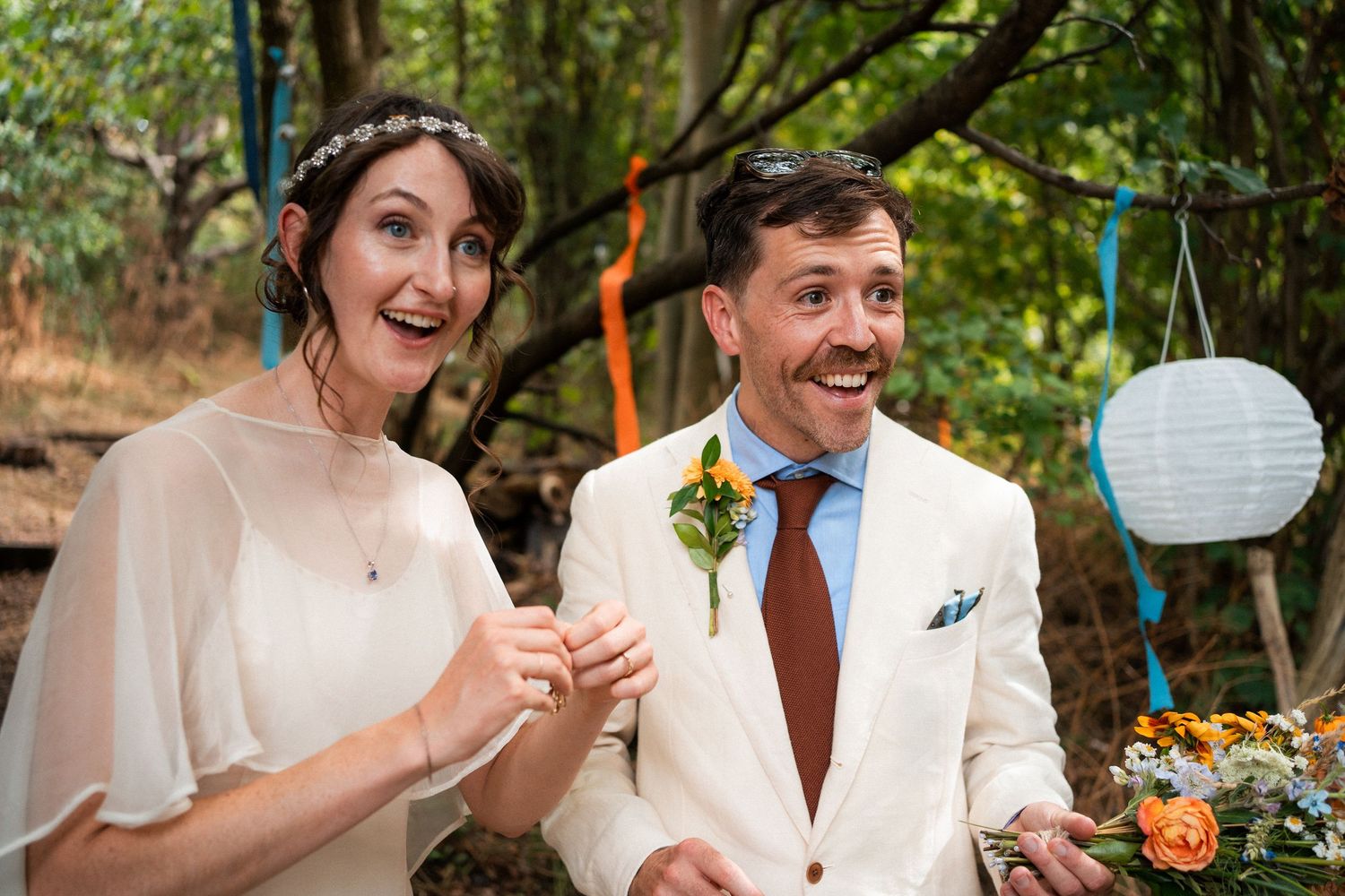 Bride and groom laughing joyfully at outdoor woodland wedding ceremony with colorful decorations and paper lanterns.