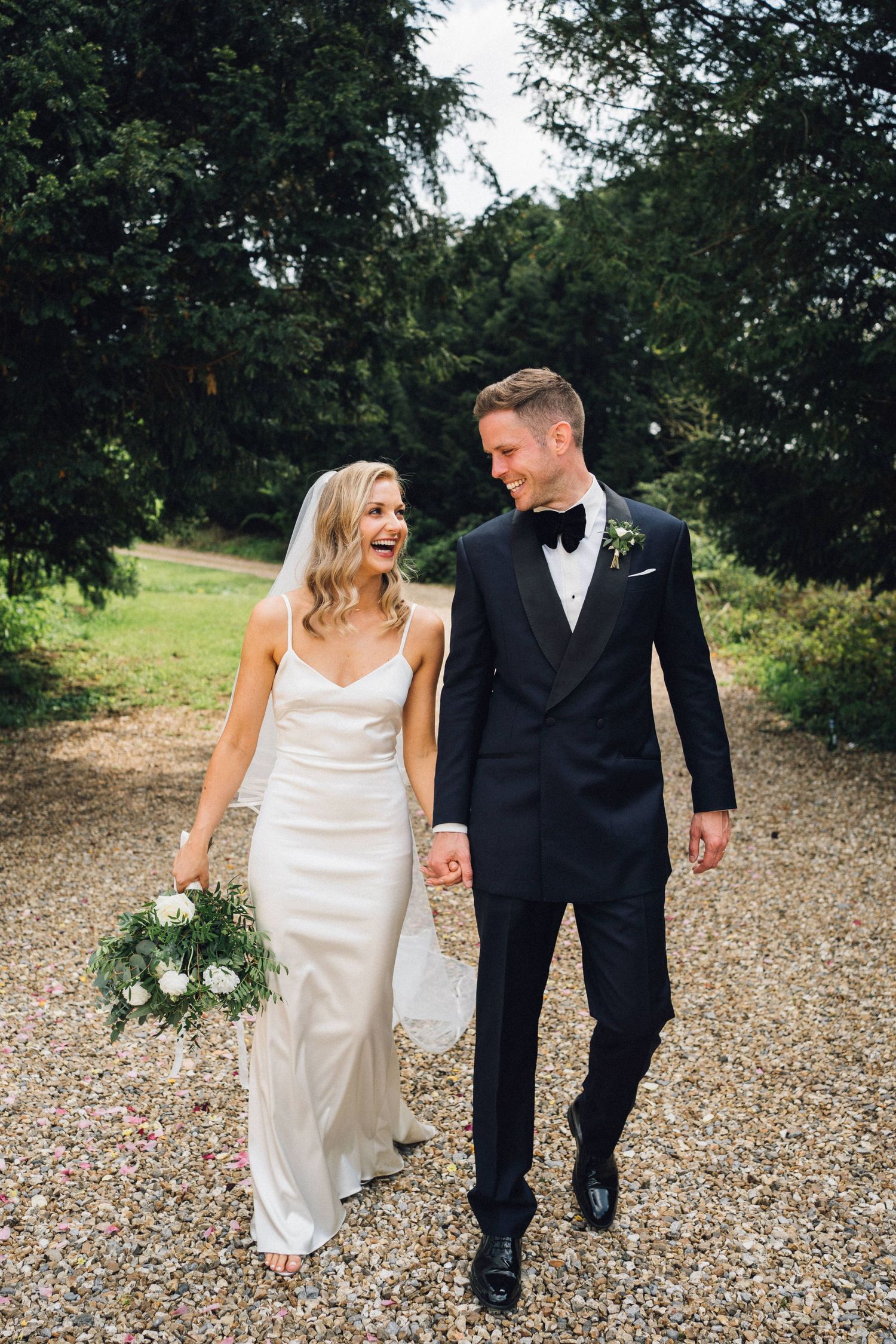 Bride and groom walking hand in hand on a gravel path surrounded by trees, smiling at each other on their wedding day.