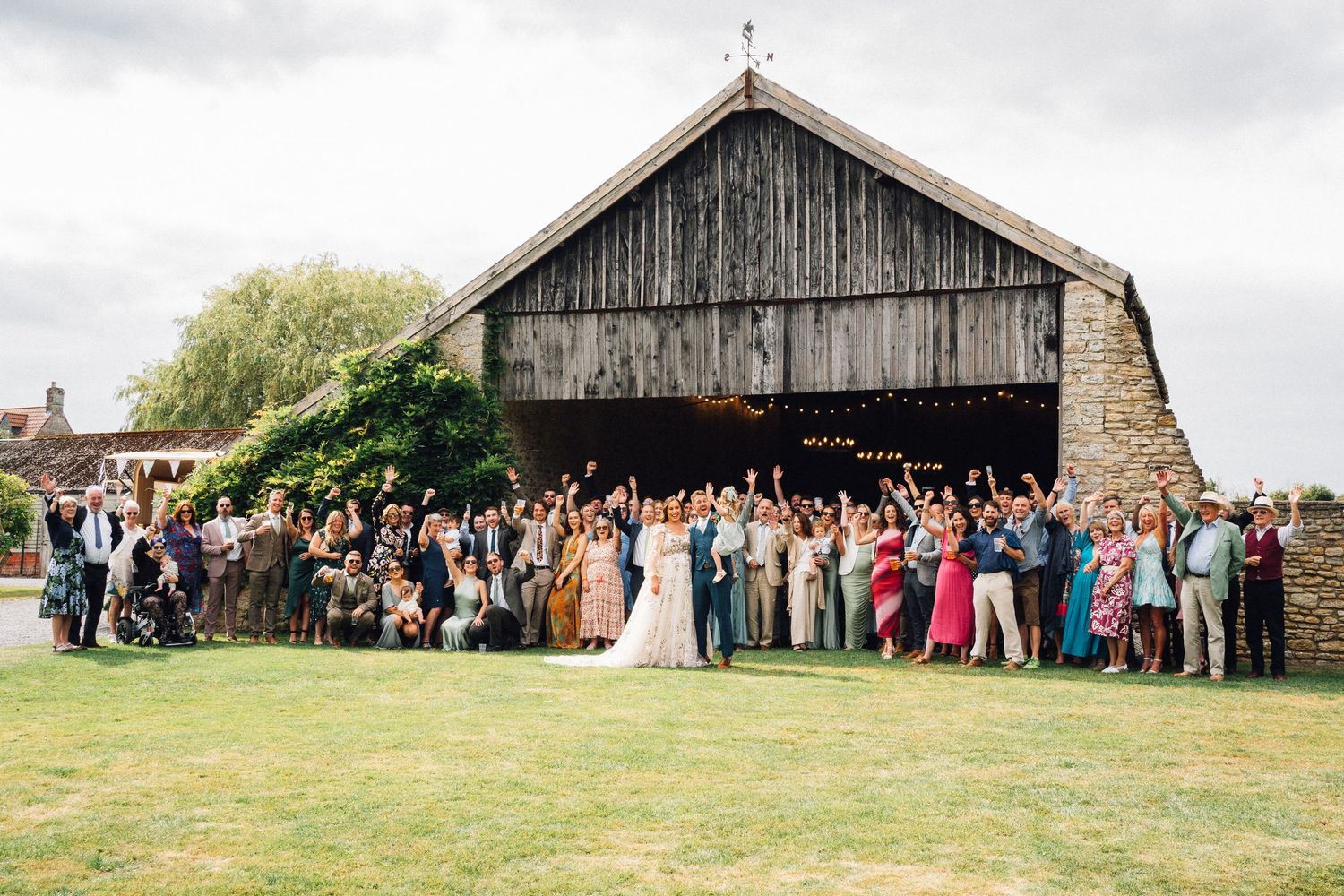 Large wedding party celebrating outside a rustic barn venue, with guests cheering on a green lawn.