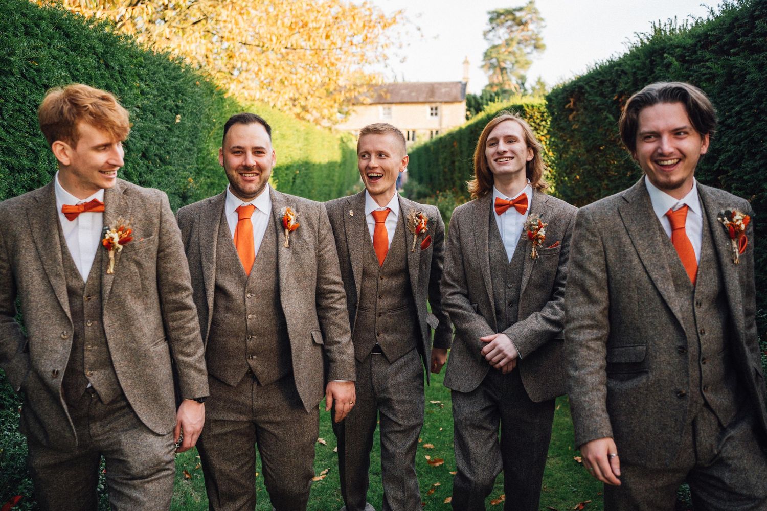 Groomsmen in brown tweed suits with orange ties pose together in a garden setting.