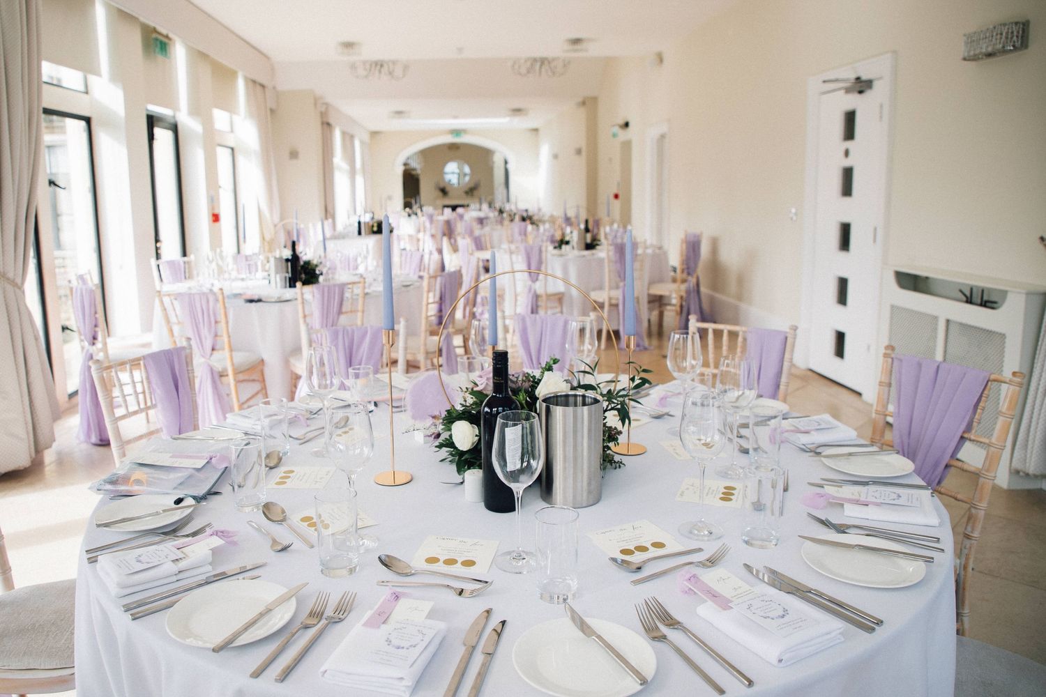 Elegant white banquet tables with formal place settings stretch down a bright modern dining room with natural lighting.