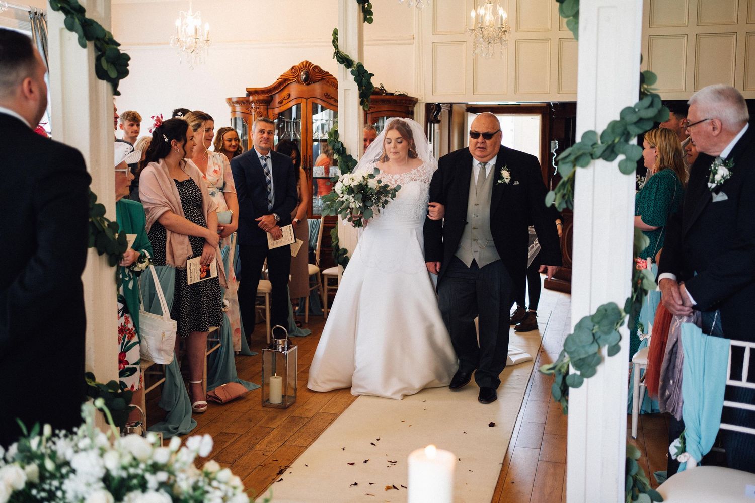 Wedding guests gather in an elegant white-walled reception venue with chandeliers and floral decorations.