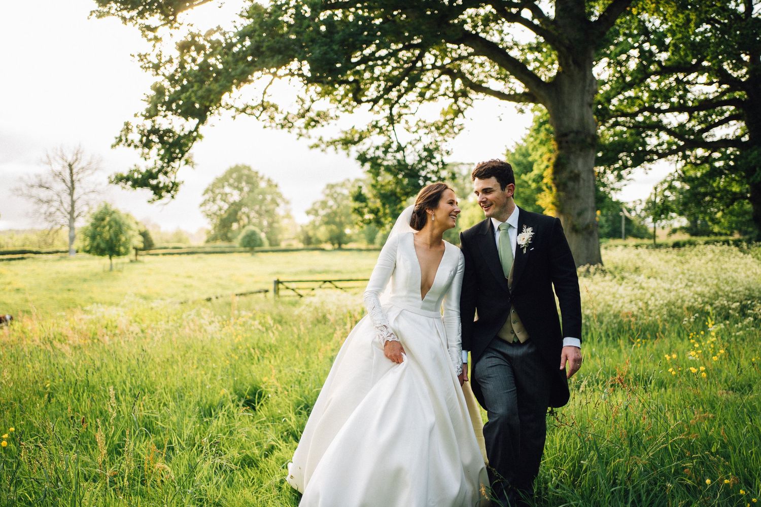 Couple walks hand in hand through sunny meadow in wedding attire.