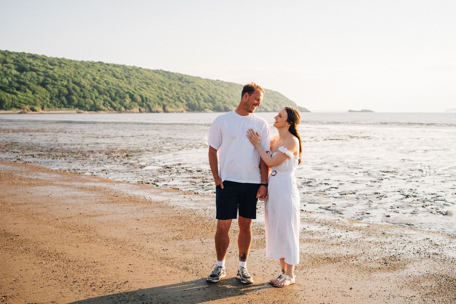 Couple in white and casual attire walking along scenic coastal beach.