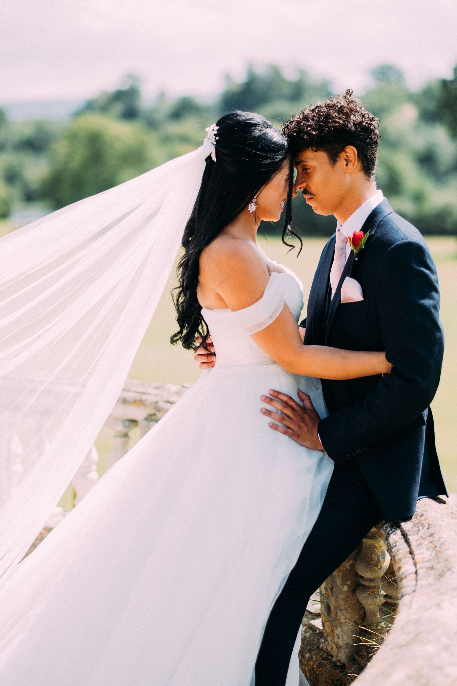 Wedding couple embracing on terrace with flowing white dress in sunlight.