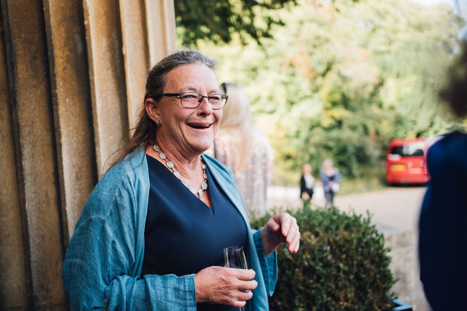 A person laughs joyfully while sitting outdoors at a restaurant table with drinks and greenery in the background.