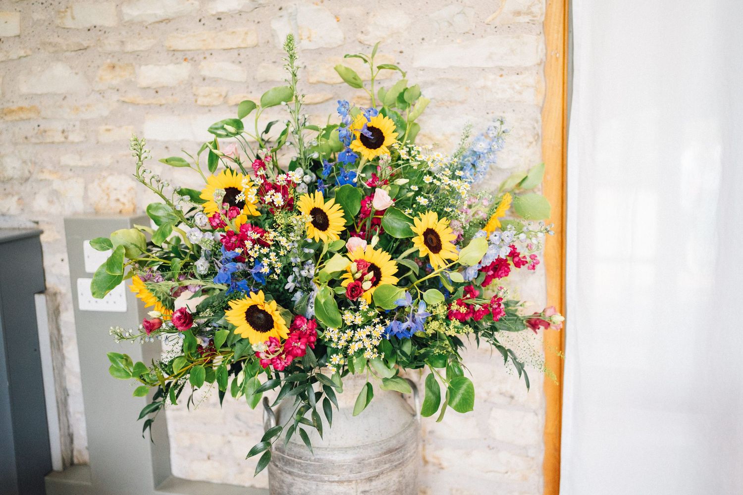 Bright floral arrangements featuring sunflowers, red and pink blooms displayed in rustic white vases against a light wall.