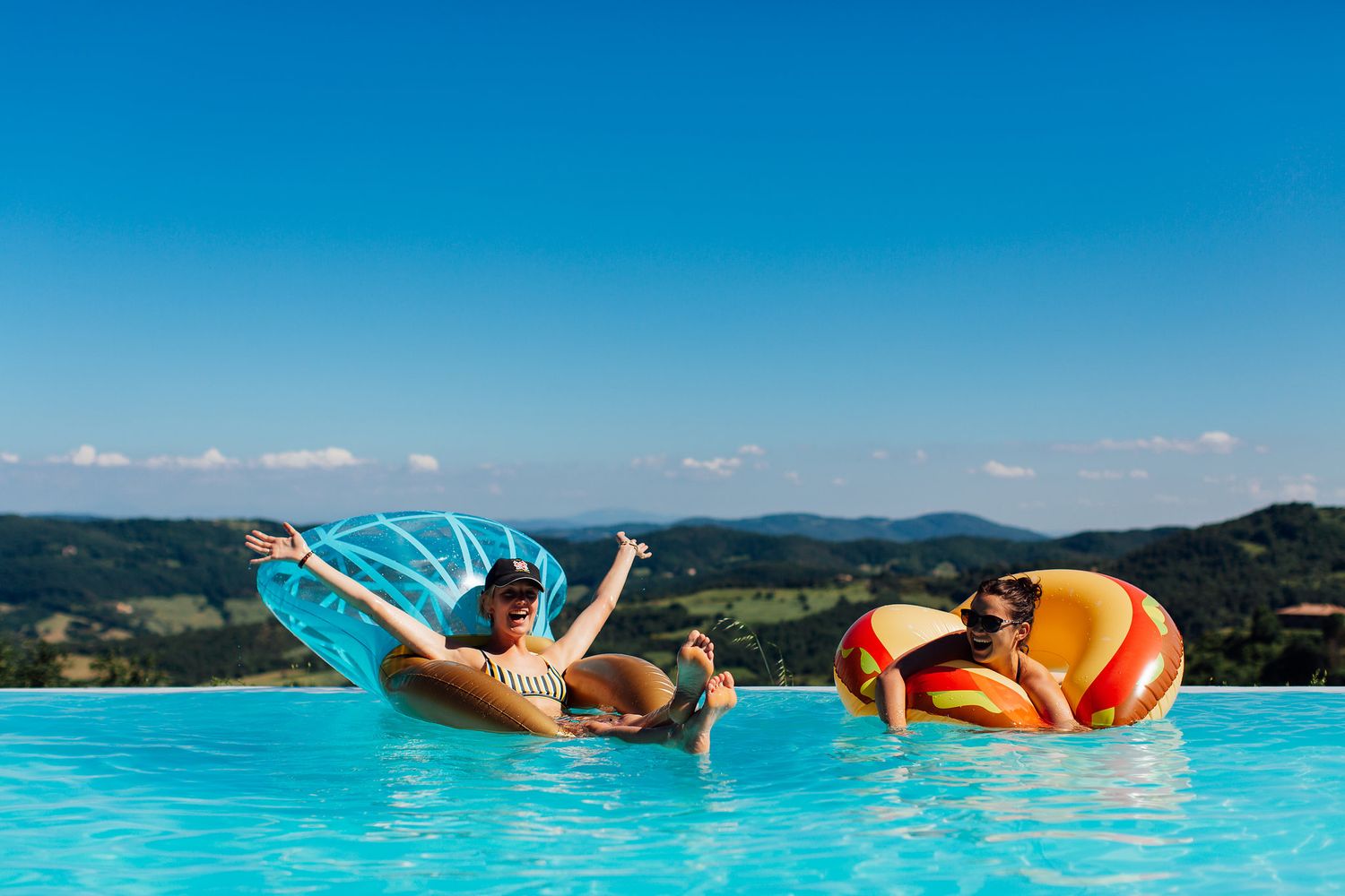 People floating and relaxing in an infinity pool overlooking a scenic tropical coastline on a sunny day.
