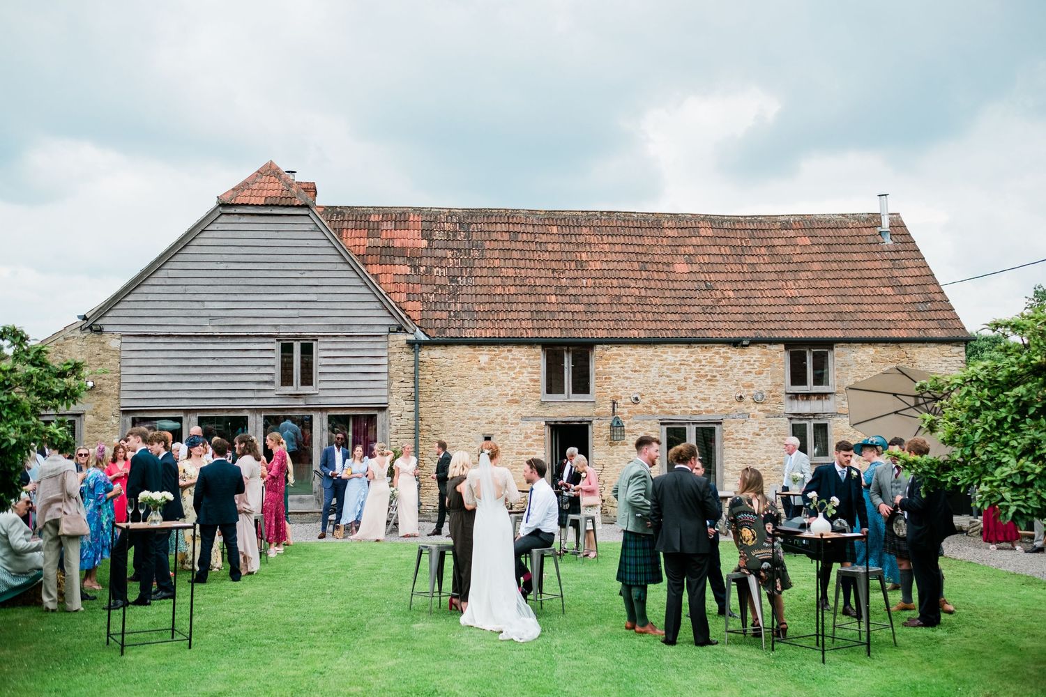 Wedding guests gather on a lawn outside a rustic barn venue with red tile roof on an overcast day.