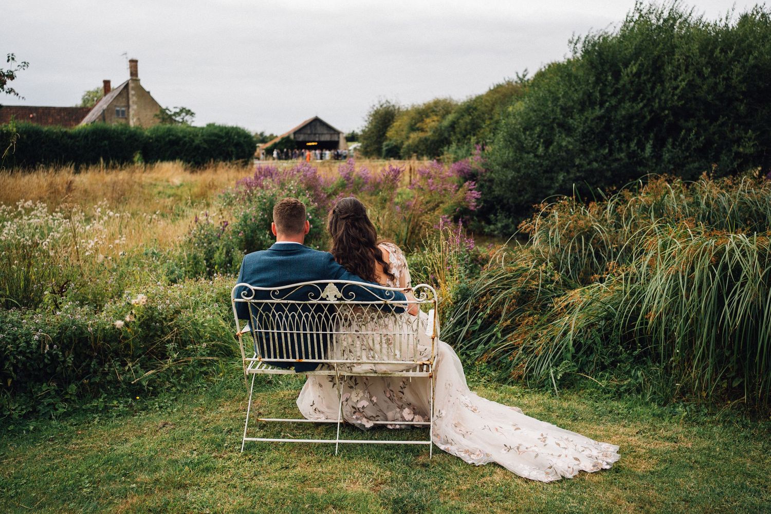 A couple sits on a wooden bench overlooking a scenic meadow on a sunny summer afternoon.