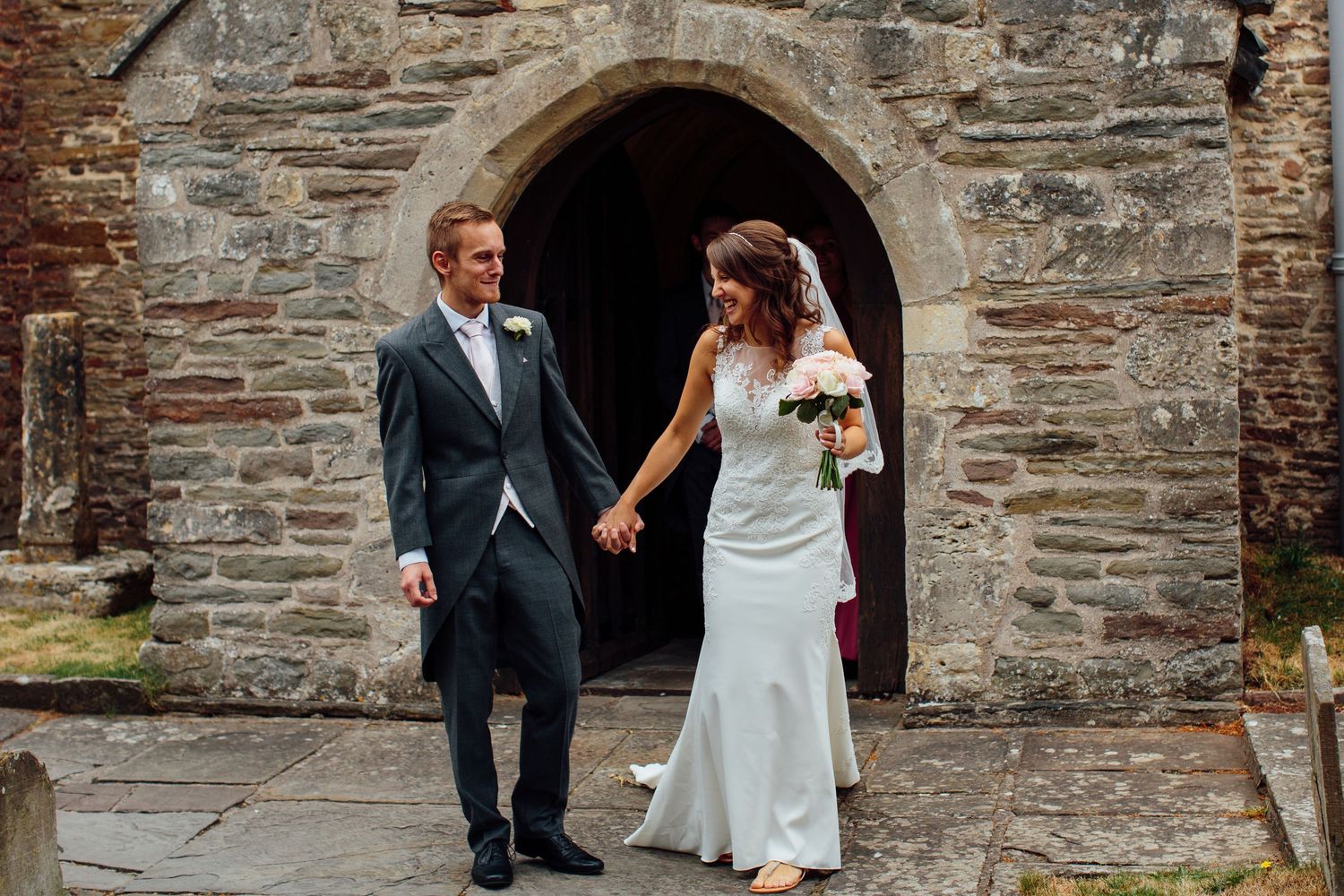 Newlyweds walk together along a rustic stone wall path outside a historic church building.