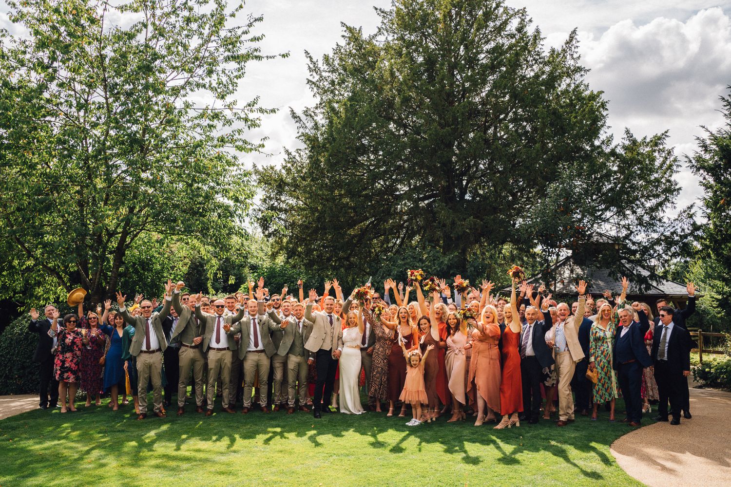 Large wedding group poses outdoors under green trees on a cloudy summer day.